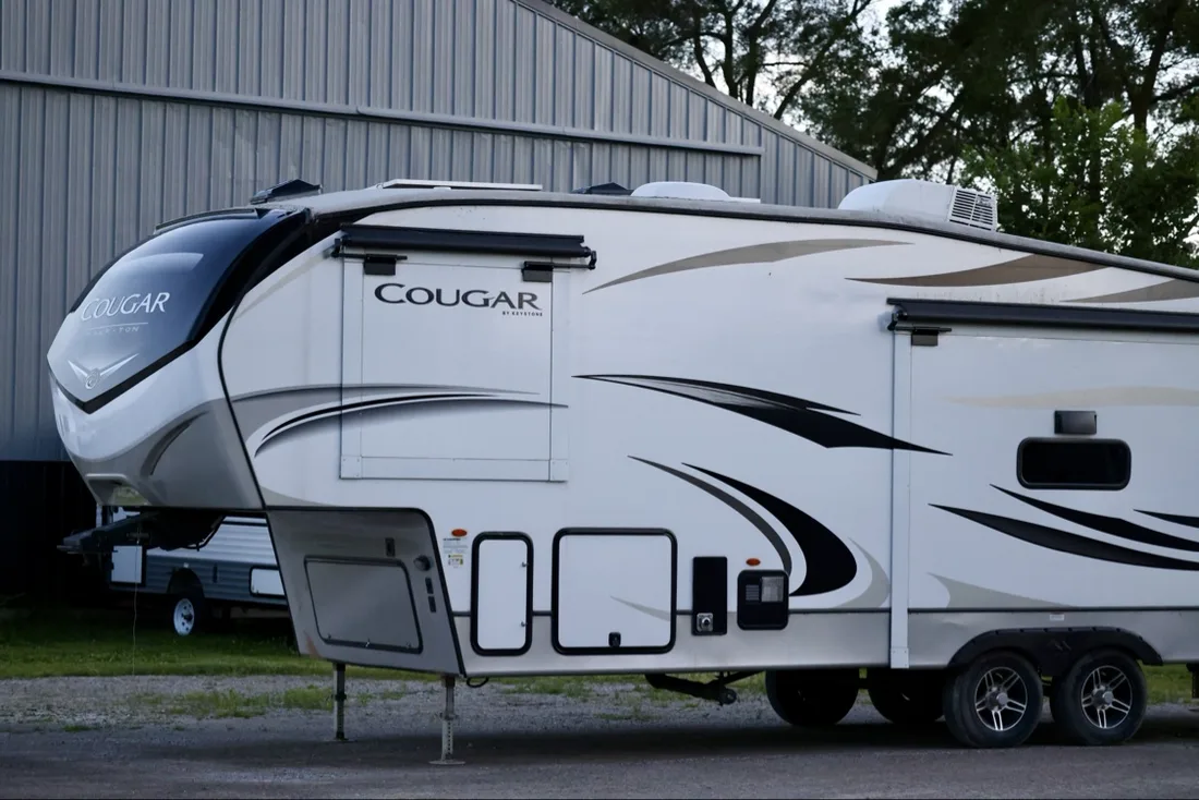 Keystone Cougar fifth wheel at the shop in Merrill, Michigan — crisp white body, wide black swoosh graphics and gold accent sweeps, the shop building behind