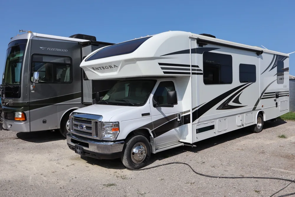 White Entegra Class C motorhome on a Ford cab with black swoosh graphics, parked beside a gray Fleetwood at Nagel Trailer Repair