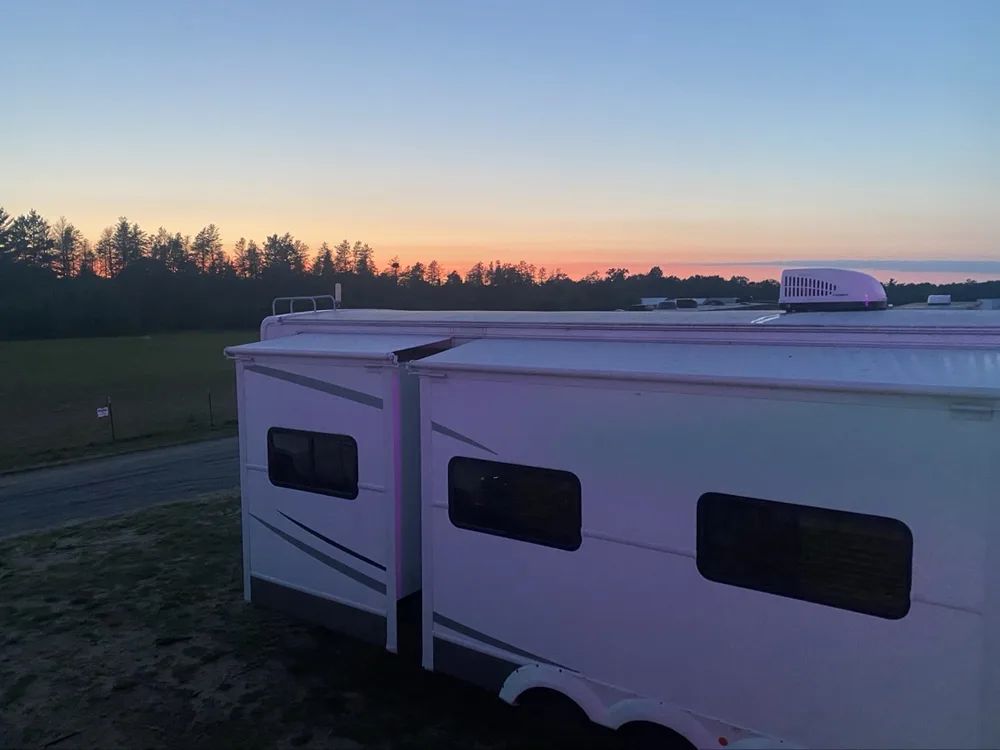 White fifth wheel with slide out photographed from roof height at sunset, pink-orange sky over the treeline at Merrill, Michigan