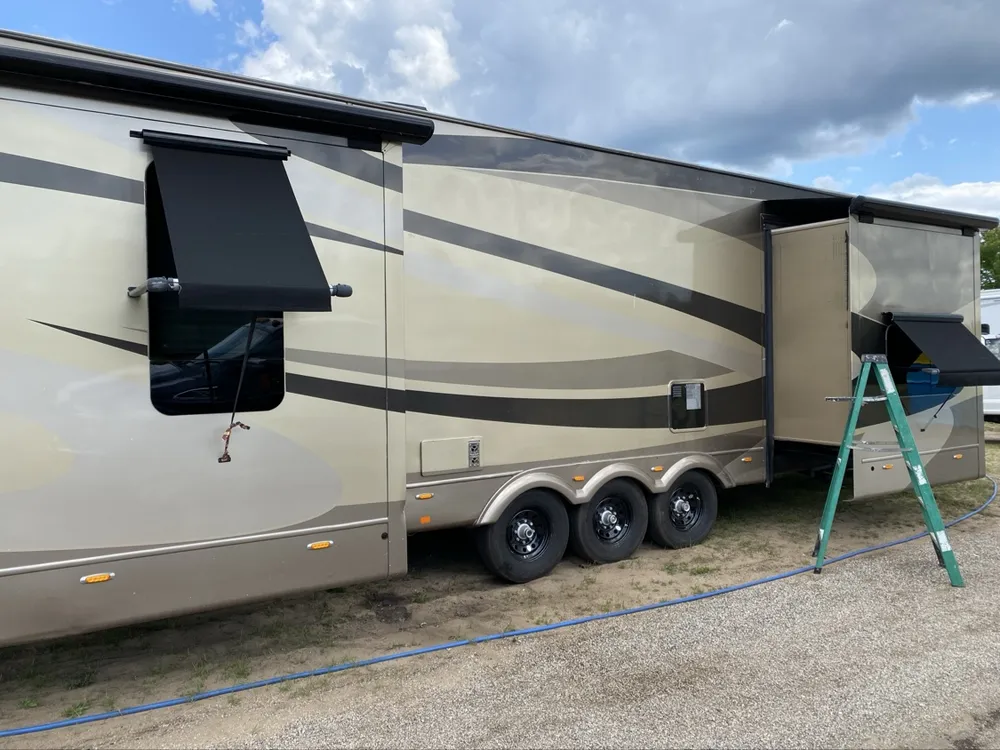 Big tan and brown triple-axle fifth wheel toy hauler with slide out and a ladder up at the slide — slide-out service in progress at Nagel Trailer Repair